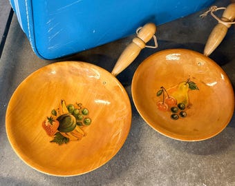 May include: Two wooden serving bowls with attached handles. The bowls are painted with a light orange hue and feature fruit illustrations. One bowl has a strawberry, banana, grapes, and a melon. The other bowl has cherries, a pear, and grapes.