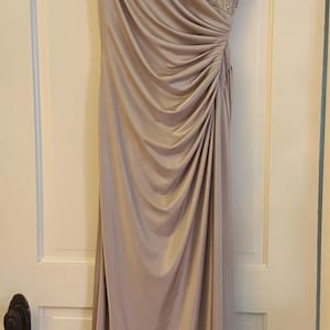 Puede incluir: Vestido de noche largo, en un suave color malva. El corpi&ntilde;o presenta intrincados detalles de cuentas, mientras que la falda drapeada a&ntilde;ade movimiento elegante. El vestido cuelga de una percha de madera contra una puerta blanca.