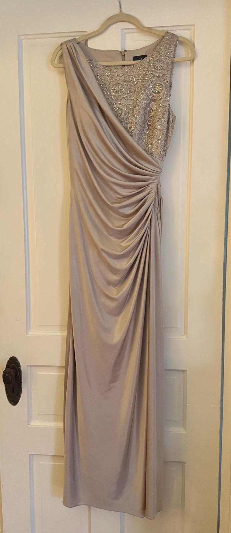 Puede incluir: Vestido de noche largo, en un suave color malva. El corpi&ntilde;o presenta intrincados detalles de cuentas, mientras que la falda drapeada a&ntilde;ade movimiento elegante. El vestido cuelga de una percha de madera contra una puerta blanca.