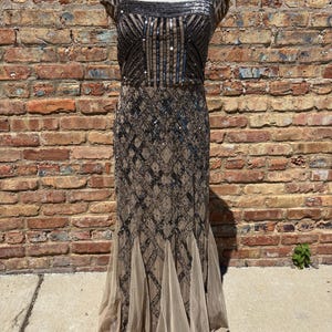 May include: A floor-length, formal dress with a square neckline and cap sleeves. The bodice and upper skirt are embellished with black sequins and beads in a geometric pattern. The lower skirt features a flowing, sheer, beige fabric.