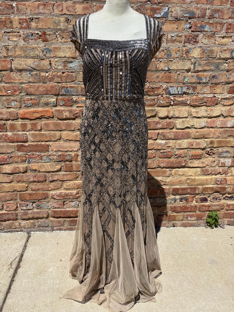 May include: A floor-length, formal dress with a square neckline and cap sleeves. The bodice and upper skirt are embellished with black sequins and beads in a geometric pattern. The lower skirt features a flowing, sheer, beige fabric.