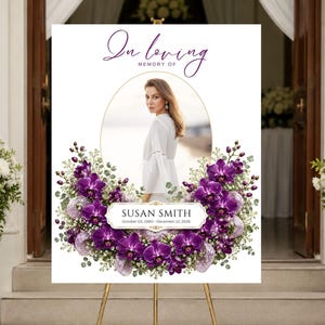 May include: A white memorial sign on a gold tripod stand featuring the text In loving MEMORY OF, a central oval photo of a person, and a nameplate reading SUSAN SMITH October 03, 1980 - December 12, 2026, surrounded by purple orchids and greenery.