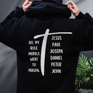 May include: Black hoodie with white text and a cross design. The text reads "ALL MY ROLE MODELS WENT TO PRISON" on the left side and lists names like Jesus, Paul, and John on the right side.