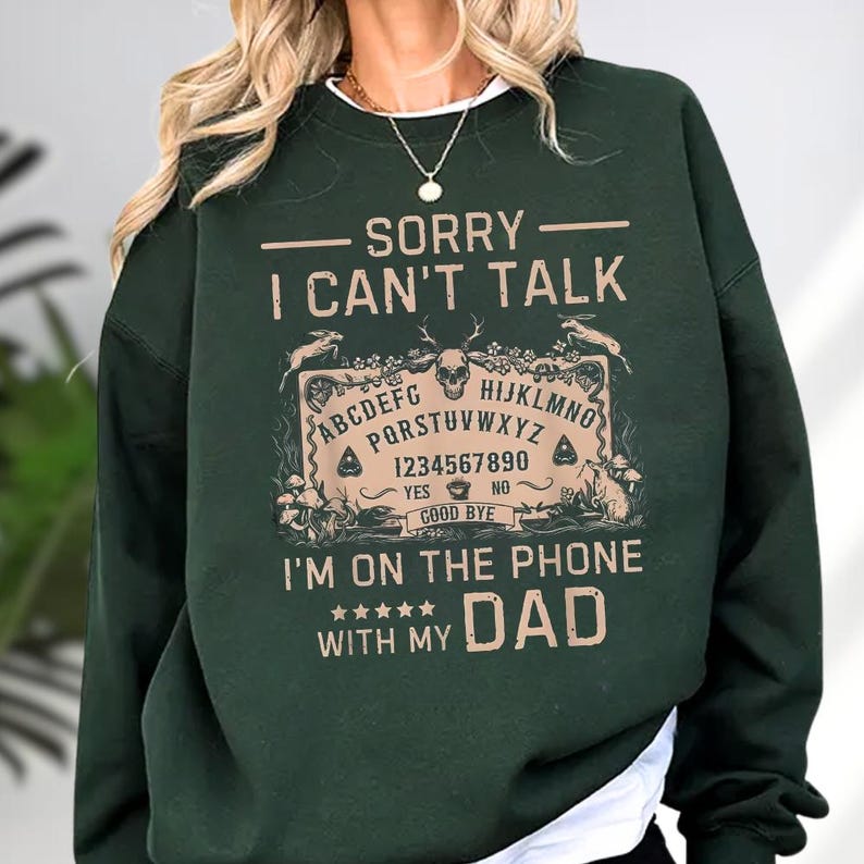 May include: Dark green sweatshirt with the text "SORRY I CAN'T TALK" above a Ouija board graphic. Below the graphic, the text reads "I'M ON THE PHONE WITH MY DAD". The design is in a tan color.