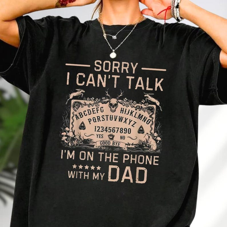 May include: Black t-shirt with the text "SORRY I CAN'T TALK" above a Ouija board graphic. Below the graphic, the text reads "I'M ON THE PHONE WITH MY DAD". The design is in a tan color.
