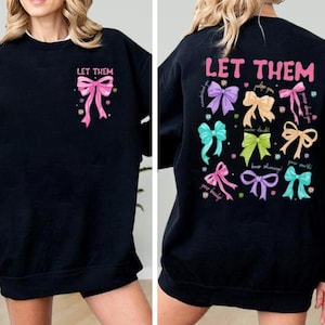 May include: Black sweatshirt with the text "LET THEM" and a pink bow graphic on the front. The back features multiple colorful bow graphics and the text "LET THEM" with inspirational phrases like "never doubt" and "keep shining!"