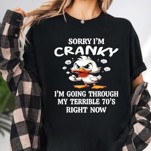 May include: Black t-shirt with the text "SORRY I'M CRANKY" and "I'M GOING THROUGH MY TERRIBLE 70'S RIGHT NOW". The shirt features a cartoon duck with an angry expression. The duck has white feathers, an orange beak, and green eyes.