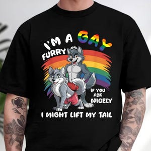 May include: A black t-shirt featuring a graphic of two grey anthropomorphic wolves wearing red shorts. Behind them is a rainbow arc. The text reads: I'M A GAY FURRY AND IF YOU ASK NICELY I MIGHT LIFT MY TAIL. The word GAY is in rainbow colors.