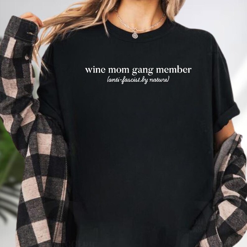 A person wearing a black t-shirt with the text "wine mom gang member (anti-fascist by nature)" printed on it.