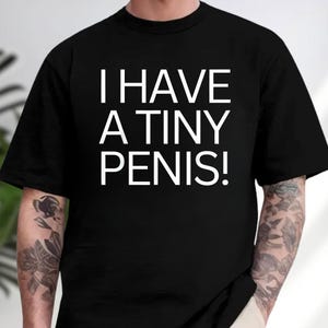 May include: Black t-shirt with the white text "I HAVE A TINY PENIS!". The shirt has short sleeves and a crew neck. The person wearing the shirt has tattoos on their arms.
