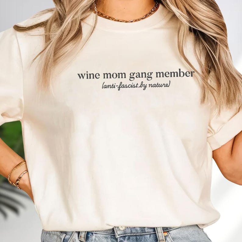 A woman wearing a white t-shirt with the text "wine mom gang member anti-fascist by nature" printed on it.