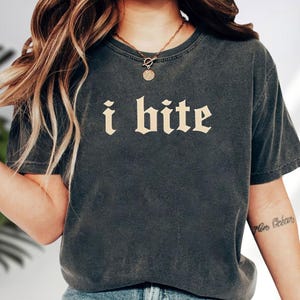 May include: Dark gray t-shirt with the text "i bite" in a gothic font. The shirt has a vintage, worn look. A gold necklace with a pendant is visible. The sleeves are short and the shirt appears to be made of a soft material.