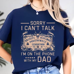 May include: Navy blue t-shirt with the text "SORRY I CAN'T TALK" above a Ouija board design. The design includes letters, numbers, and the words "YES" and "NO". Below the design is the text "I'M ON THE PHONE WITH MY DAD".