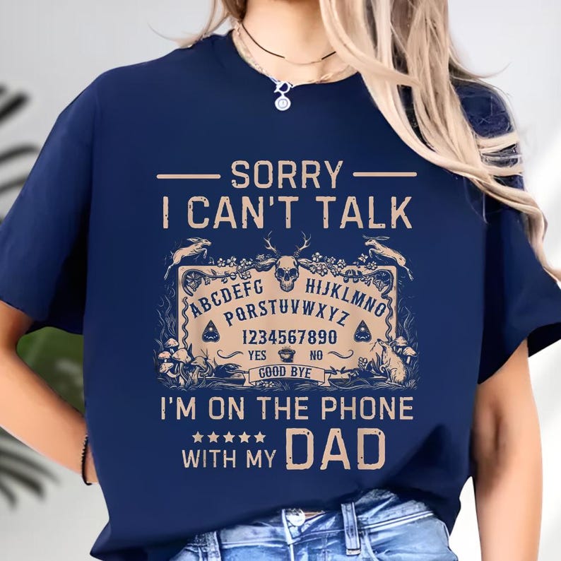 May include: Navy blue t-shirt with the text "SORRY I CAN'T TALK" above a Ouija board design. The design includes letters, numbers, and the words "YES" and "NO". Below the design is the text "I'M ON THE PHONE WITH MY DAD".