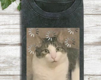 May include: Dark gray t-shirt featuring a distressed graphic of a cat with loading symbols around its head. The cat has white and gray fur and a surprised expression. The shirt is folded on a light wood surface.