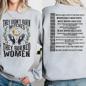 May include: A light gray sweatshirt featuring a front graphic with two hands, a crescent moon, and the text "THEY DIDN'T BURN WITCHES THEY BURNED WOMEN." The back displays a timeline of historical dates and text regarding women's rights milestones.