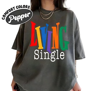 May include: Dark grey t-shirt with the words "LIVING Single" in colorful block letters. The word "LIVING" is in red, yellow, blue, purple, and orange. The shirt has the text "COMFORT COLORS Pepper" in a black oval.