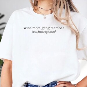 A person wearing a white t-shirt with the text "wine mom gang member [anti-fascist by nature]" printed on it.