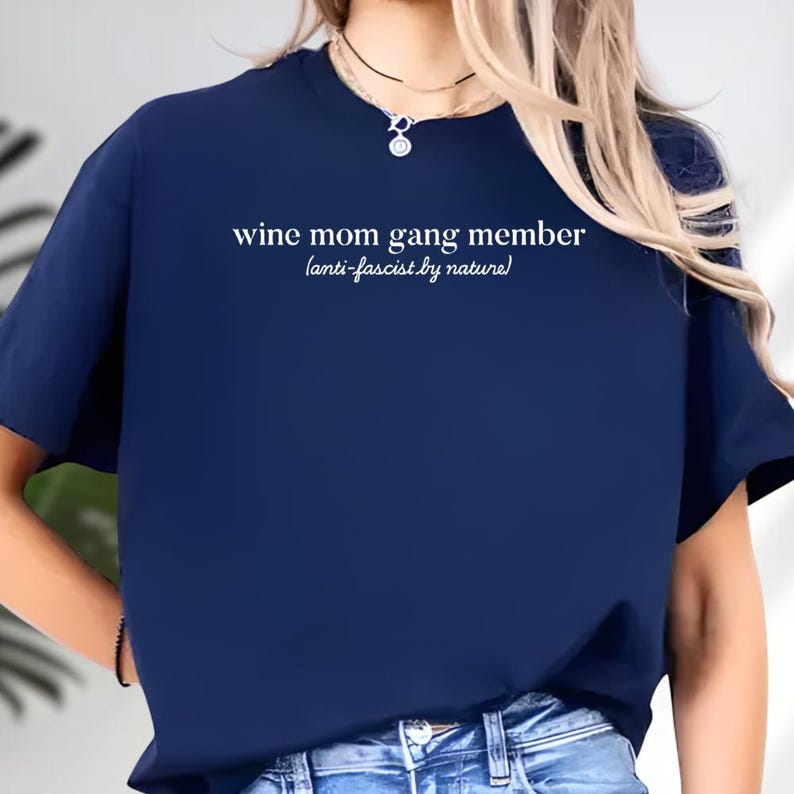 A person wearing a navy blue t-shirt with the text "wine mom gang member anti-fascist by nature!" printed on it.