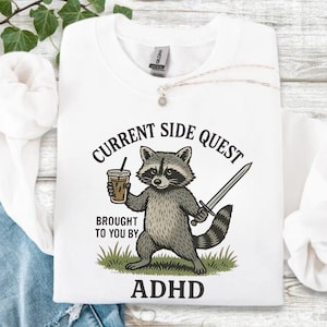 May include: White sweatshirt with a graphic of a raccoon holding a coffee and a sword. Text reads "CURRENT SIDE QUEST BROUGHT TO YOU BY ADHD". The raccoon is gray and black, standing on green grass.