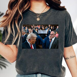 May include: Dark gray t-shirt with a printed image of a crowd of people, including Donald Trump, and a sign that reads "BLACK PEOPLE AREN'T APES!". The shirt has a crew neck and short sleeves.
