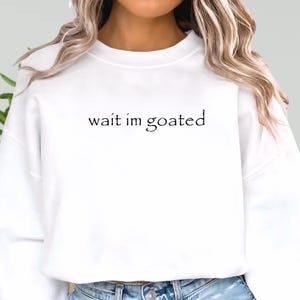 a woman wearing a white sweatshirt that says wait im goated