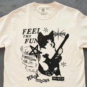 May include: Cream-colored t-shirt with a black graphic design. The design features the text "FEEL THE FUN" and "MEGA CYCLONE", along with a cat playing a guitar and other symbols. The shirt has a crew neck and short sleeves.