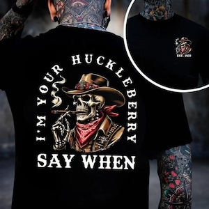May include: Black t-shirt featuring a skull wearing a cowboy hat, red bandana, and smoking a cigar. The text reads "I'M YOUR HUCKLEBERRY SAY WHEN." A smaller image of the skull with "EST. 1972" is on the upper right.