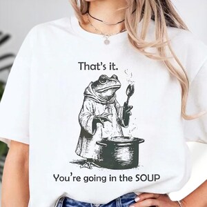 May include: White t-shirt with a black and white illustration of a frog wearing a robe and glasses, holding a spoon over a pot. The text "That's it." is above the frog, and "You're going in the SOUP" is below.