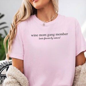 A woman wearing a pink t-shirt with the text "wine mom gang member [antislaverybynatural]" printed on it.