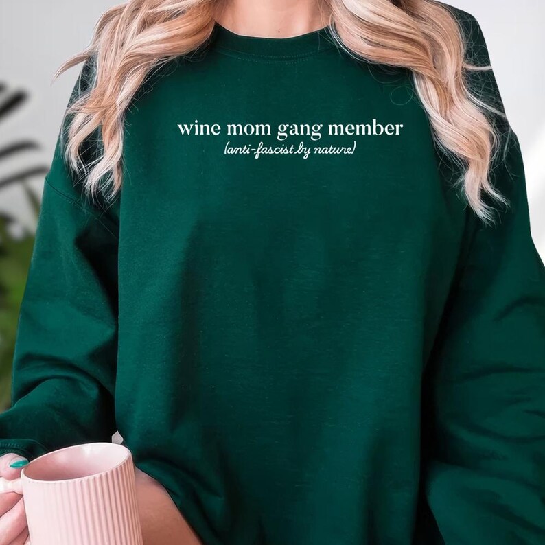 A person wearing a green sweatshirt with the text "wine mom gang member [anti-fascist by nature]" printed on it.