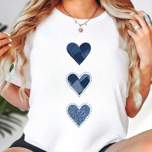 May include: A white short-sleeved t-shirt featuring three vertically aligned heart graphics. Each heart displays a different blue denim-inspired pattern, including patchwork textures and a speckled wash, all outlined with a dashed stitch effect.