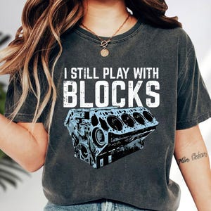 May include: Dark gray t-shirt with the text "I STILL PLAY WITH BLOCKS" above a light blue engine block graphic. The shirt is a casual style, suitable for everyday wear.