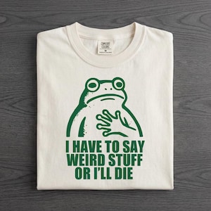 May include: An off-white crew neck t-shirt featuring a dark green graphic of a frog with one hand placed on its chest. Below the frog, the text reads: I HAVE TO SAY WEIRD STUFF OR I'LL DIE. The shirt has a Comfort Colors brand label at the neck.