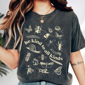 May include: Dark gray t-shirt featuring a variety of insect illustrations in a cream color. The shirt has the text "be kind to all kinds" in a circular design. The shirt is a crew neck and has short sleeves.