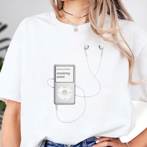 May include: White t-shirt featuring a graphic of a gray MP3 player with the text "NOW PLAYING mommy asmr." The MP3 player has white earbuds connected. The shirt is a casual, everyday wear item.