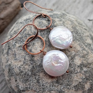 May include: A pair of handmade earrings featuring large, lustrous white coin pearls suspended from copper-colored hoops and ear wires. The earrings are displayed on a gray stone surface, showcasing their natural beauty and unique design.