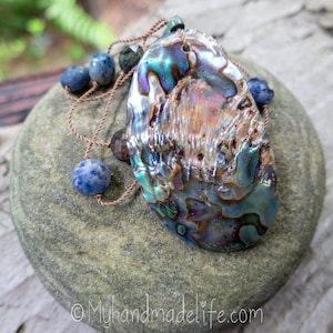 May include: An iridescent abalone shell pendant necklace with blue and brown gemstone beads. The shell displays a range of colors, including green, blue, and purple. The necklace is strung on a brown cord. The website address myhandmadelife.com is visible.