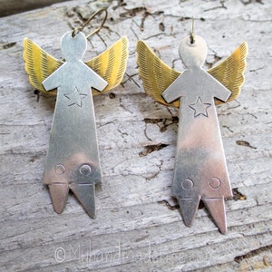 May include: A pair of handmade angel earrings. Each earring has a silver-coloured body with a star detail and gold-coloured wings. The earrings are displayed on a weathered wooden surface.