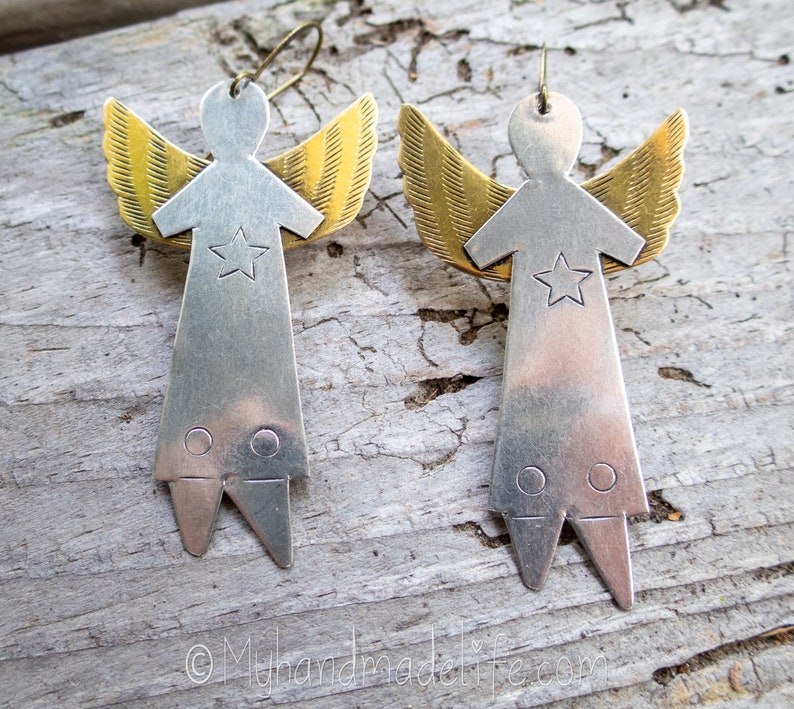 May include: A pair of handmade angel earrings. Each earring has a silver-coloured body with a star detail and gold-coloured wings. The earrings are displayed on a weathered wooden surface.
