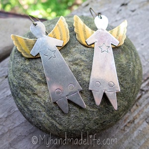 May include: A pair of handmade angel earrings. Each earring has a silver-coloured body with a star cutout and gold-coloured wings. The earrings are displayed on a grey stone surface.