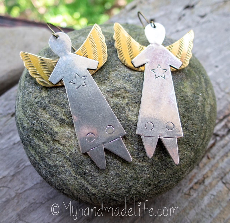 May include: A pair of handmade angel earrings. Each earring has a silver-coloured body with a star cutout and gold-coloured wings. The earrings are displayed on a grey stone surface.