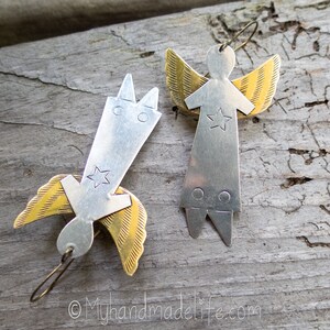 May include: Two handmade angel earrings. Each earring features a silver body with a star and gold wings. One angel is upright, the other is inverted. The earrings are on a wooden surface.