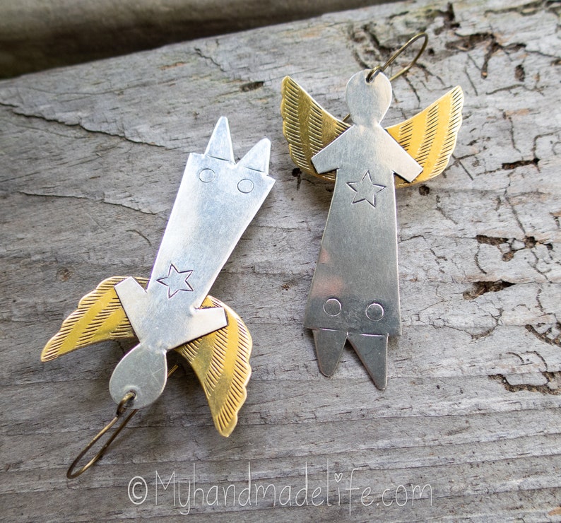 May include: Two handmade angel earrings. Each earring features a silver body with a star and gold wings. One angel is upright, the other is inverted. The earrings are on a wooden surface.