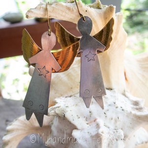 May include: Handmade angel earrings with a star design. The earrings feature a silver body, brass wings, and a copper-coloured torso. The earrings are hanging from gold-coloured hooks. The earrings are displayed on a white seashell.