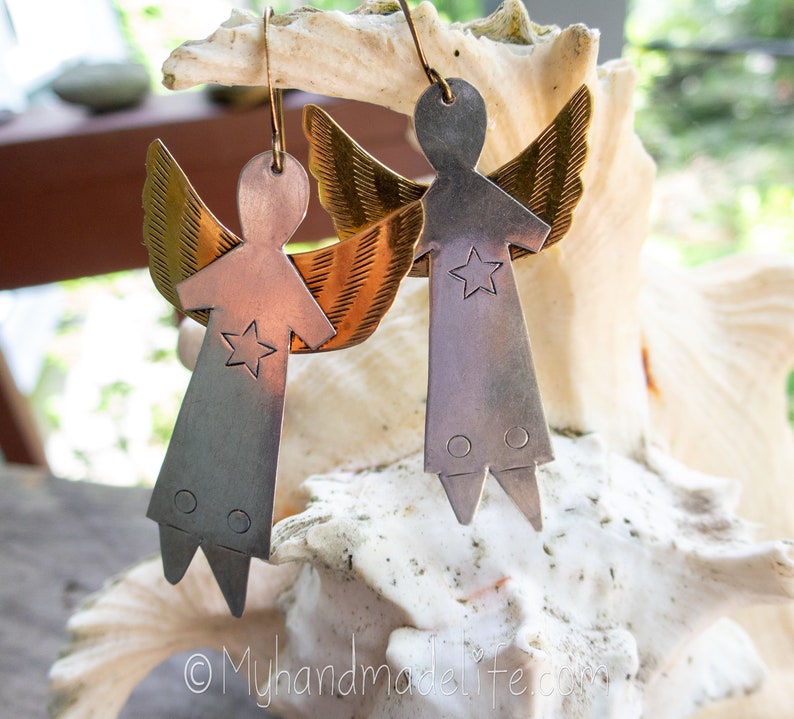 May include: Handmade angel earrings with a star design. The earrings feature a silver body, brass wings, and a copper-coloured torso. The earrings are hanging from gold-coloured hooks. The earrings are displayed on a white seashell.
