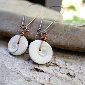 May include: Handmade earrings featuring white, spiral-shaped pendants. The pendants are attached to copper wire and silver-colored ear wires. The earrings are displayed on a natural stone surface, showcasing their organic design and unique craftsmanship.
