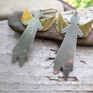 May include: A pair of angel-shaped earrings. Each earring has a silver-coloured metal body with a star and brass-coloured wings. The earrings have a simple, geometric design and are suspended from a hook.