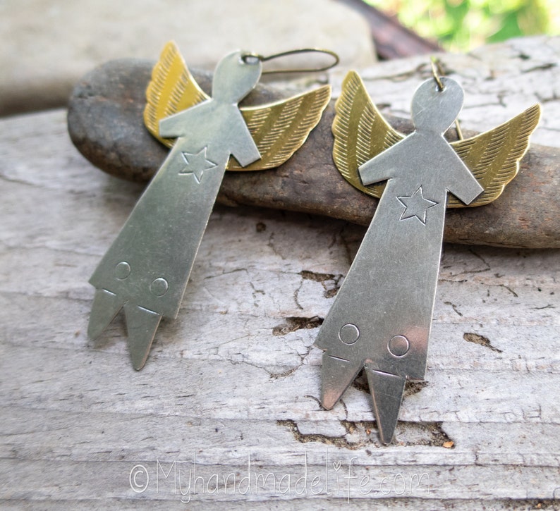 May include: A pair of angel-shaped earrings. Each earring has a silver-coloured metal body with a star and brass-coloured wings. The earrings have a simple, geometric design and are suspended from a hook.