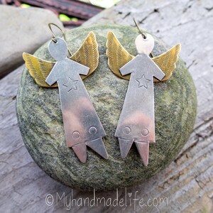May include: A pair of handmade angel earrings. Each earring has a silver-coloured body with a star and gold-coloured wings. The earrings are displayed on a grey stone, with a wooden background. The earrings have a hook closure.